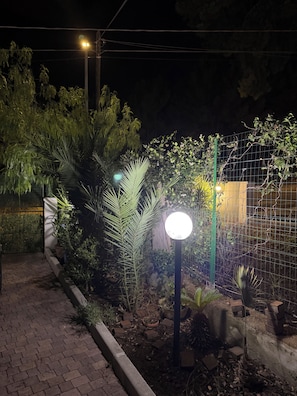 A nighttime garden with a paved walkway, various plants, and a fence. - Tereskova (Castellaneta Marina)