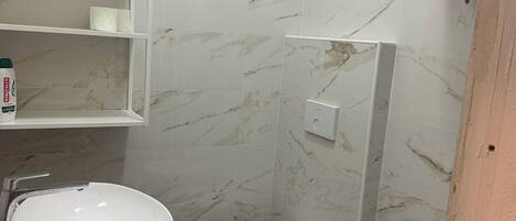 A bathroom with a white sink, toilet, and bidet, marble walls, and a shelf with toiletries.