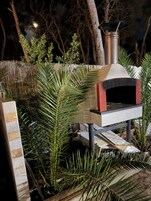 Un horno de pizza de ladrillo al aire libre con chimenea, rodeado de follaje y una valla de madera.