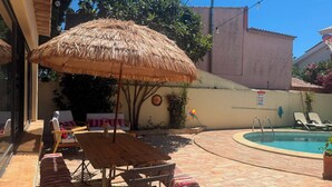 A patio with a pool, a thatched umbrella, lounge chairs, and a wooden table. - SEAS THE DAY 2 (Luz)