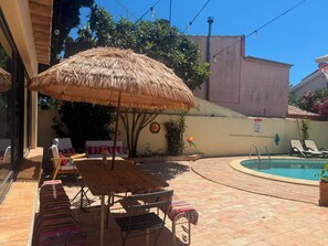 Eine Terrasse mit Pool, einem Reetdachschirm, Liegestühlen und einem Holztisch.