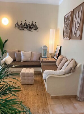 Ein Wohnzimmer mit Ecksofa, einem kleinen Hocker, einem Beistelltisch mit Lampe sowie Wandbildern.