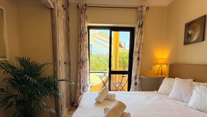 A hotel room with a bed, a potted plant, a balcony with a table and chairs, and a lamp. - SEAS THE DAY 2 (Luz)