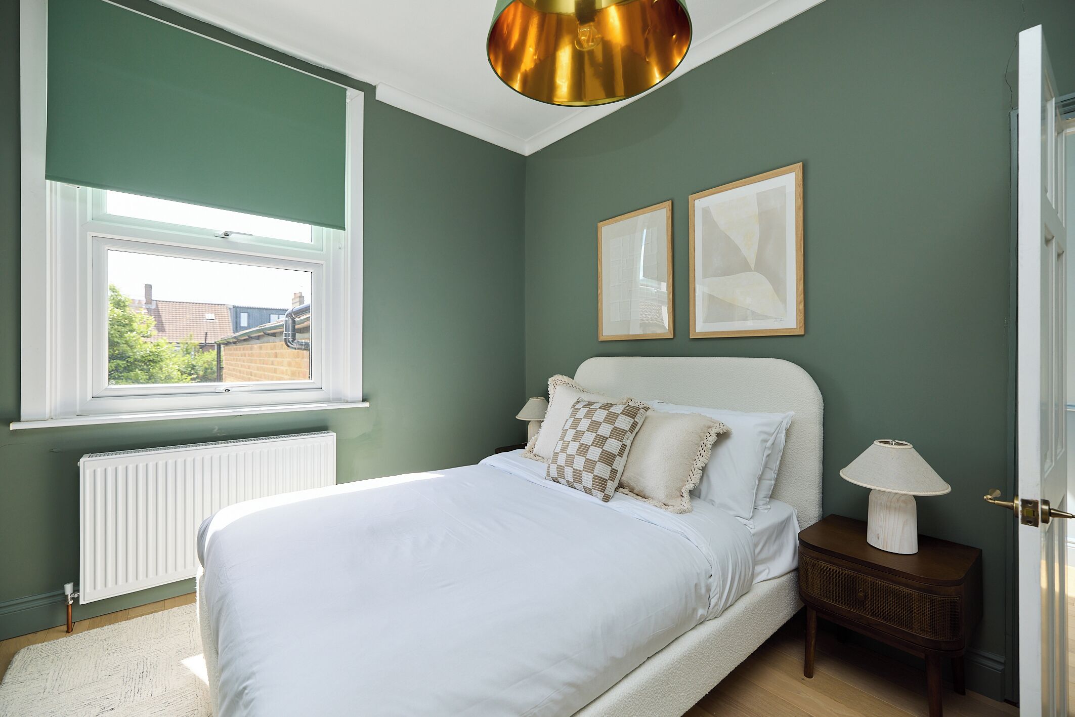 Une chambre à coucher avec un lit, une table de chevet, une lampe, une fenêtre et deux tableaux encadrés accrochés au mur.