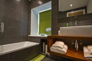 Une salle de bain moderne avec une baignoire, un mur d’accent vert, un miroir et des serviettes soigneusement pliées sur une étagère en bois.