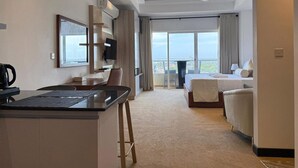A hotel room with a kitchenette, a bed, a sofa, and a large window offering a view.