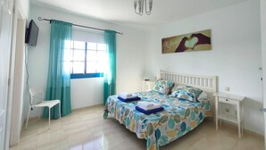 Een slaapkamer met een bed, een stoel, een televisie, een schilderij en een raam met gordijnen.