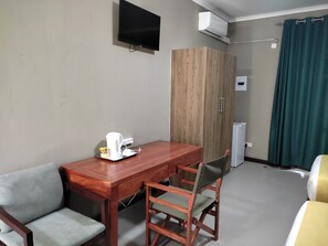 A hotel room with a wooden table, two chairs, a television, an air conditioning unit, a wardrobe, and a bed with a green curtain.