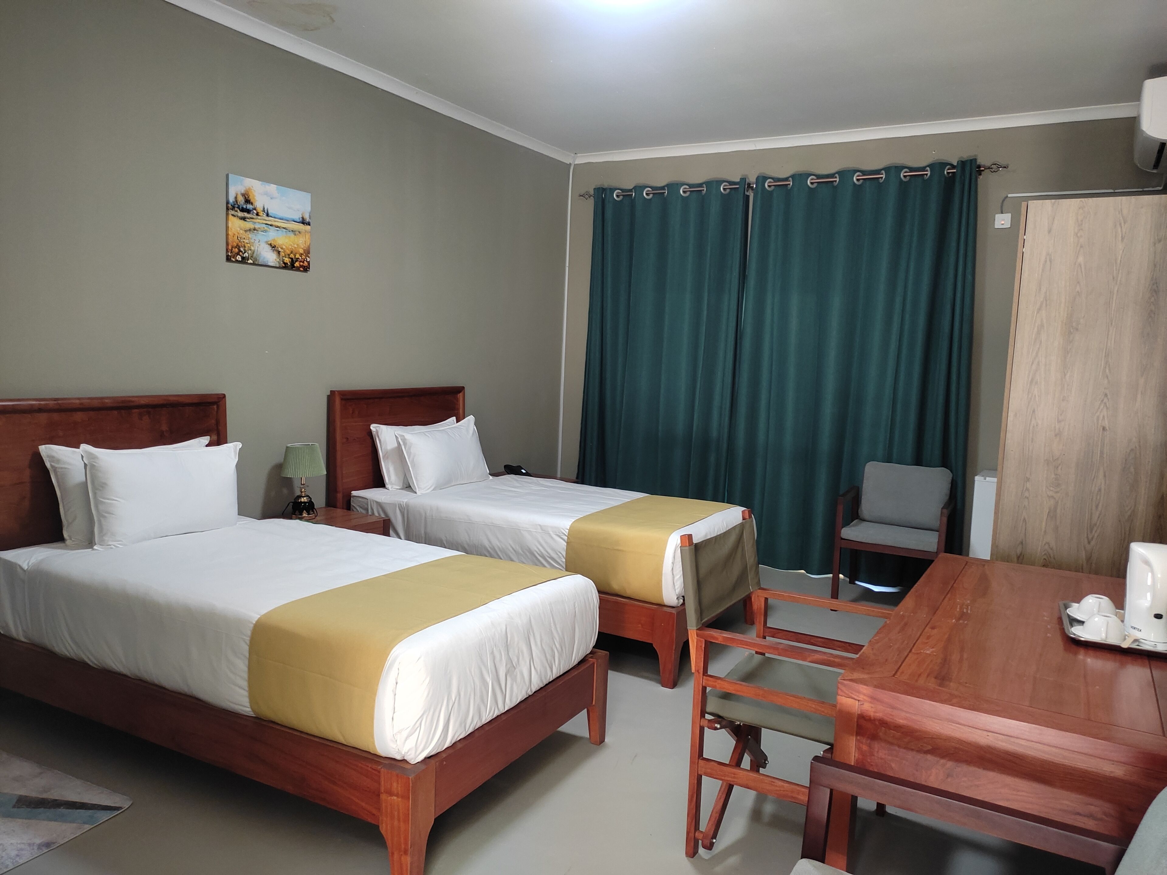 A hotel room with two beds, a wooden desk, a chair, a table with a kettle, and a painting on the wall.