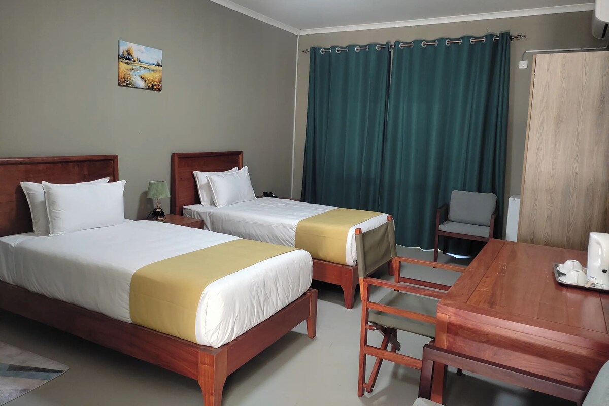 A hotel room with two beds, a wooden desk, a chair, and a painting on the wall.