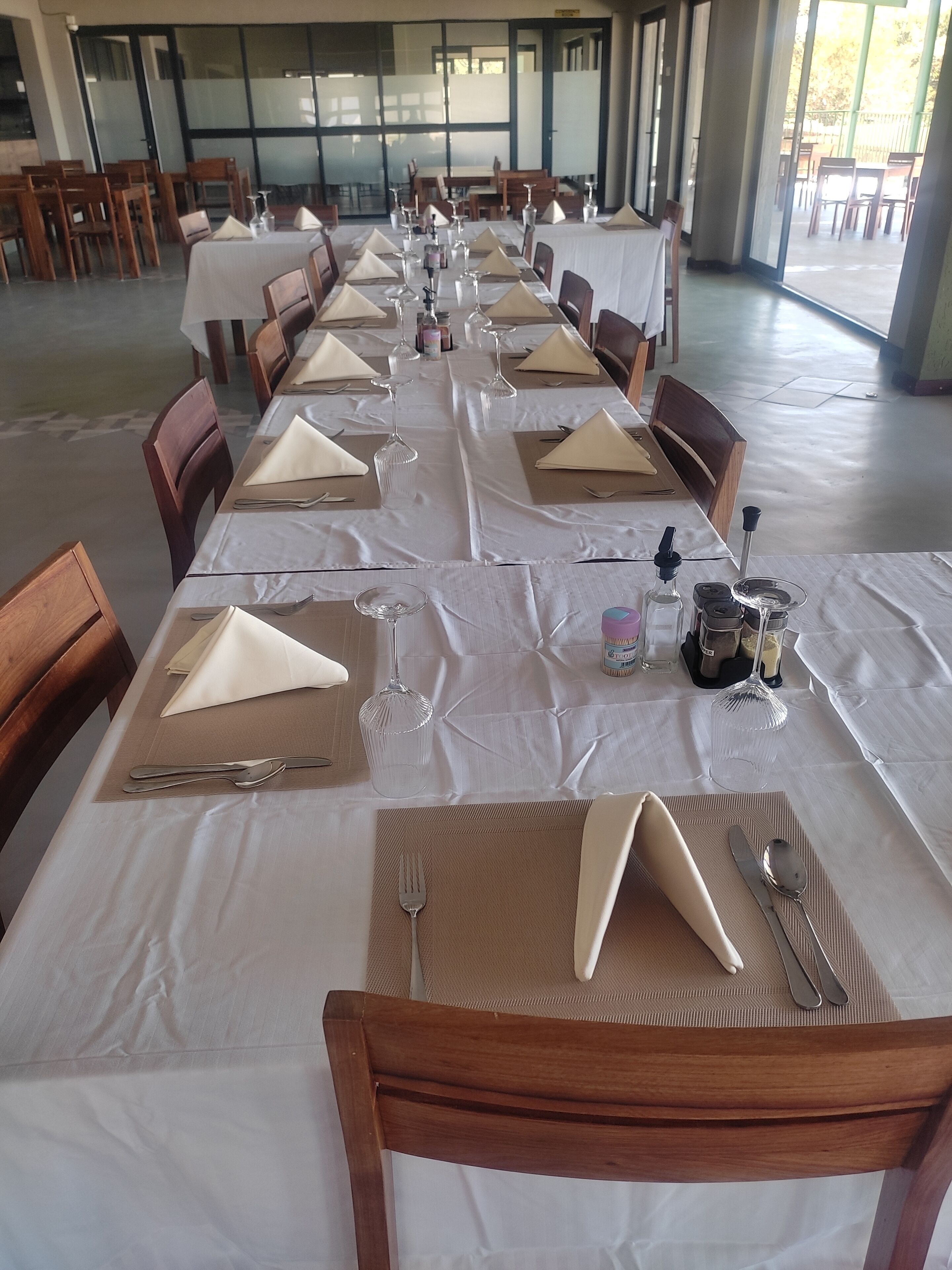 Una larga mesa de comedor dispuesta para una comida, con varias sillas, mantel blanco y cubertería y cristalería ordenadas.