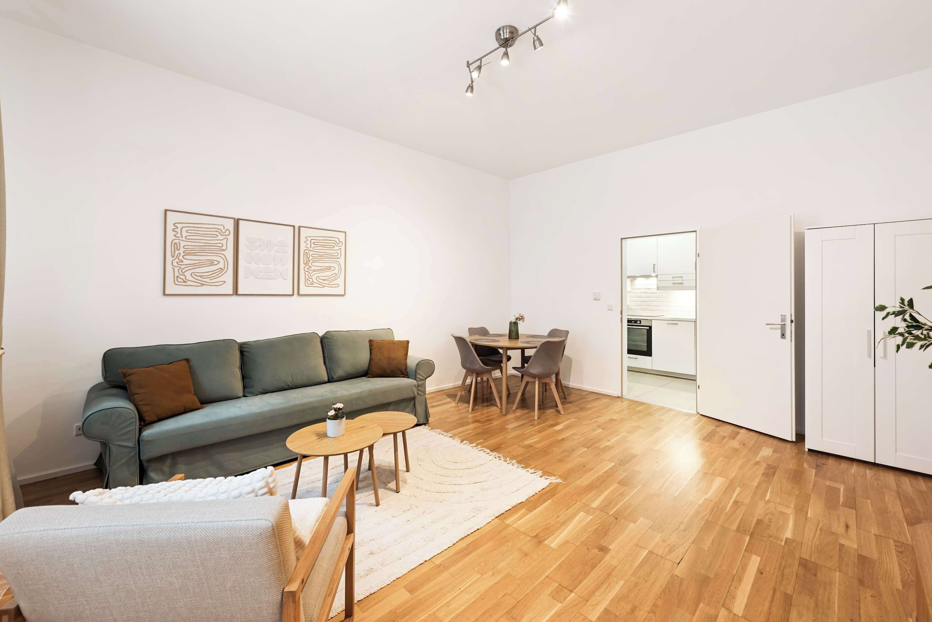 Uma sala moderna com sofá, uma pequena mesa redonda, um espaço de refeições com cadeiras e uma cozinha visível por uma porta aberta.
