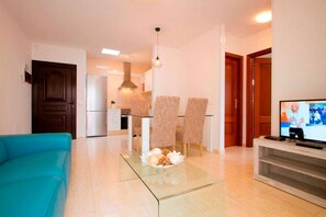 Una sala de estar moderna con una mesa de centro de vidrio, un sofá azul, un televisor y una cocina al fondo.