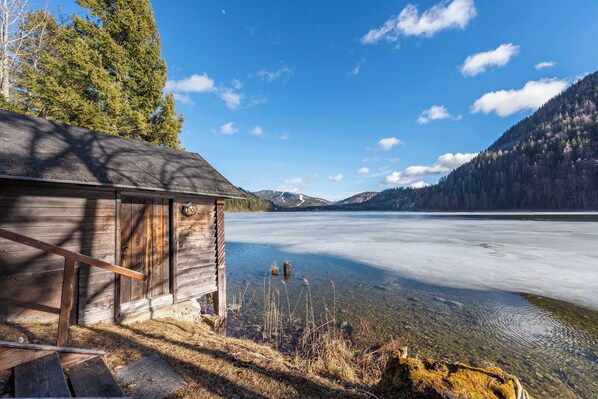 Une cabane en bois au bord d’un lac gelé aux eaux cristallines, entourée d’arbres et de montagnes.