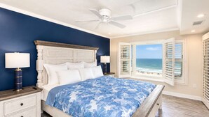 Un dormitorio con cama, mesitas de noche, ventilador de techo, ventana con vista al mar y suelo de madera.