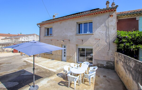 Une maison avec un patio, des chaises blanches, une table et un parasol bleu.