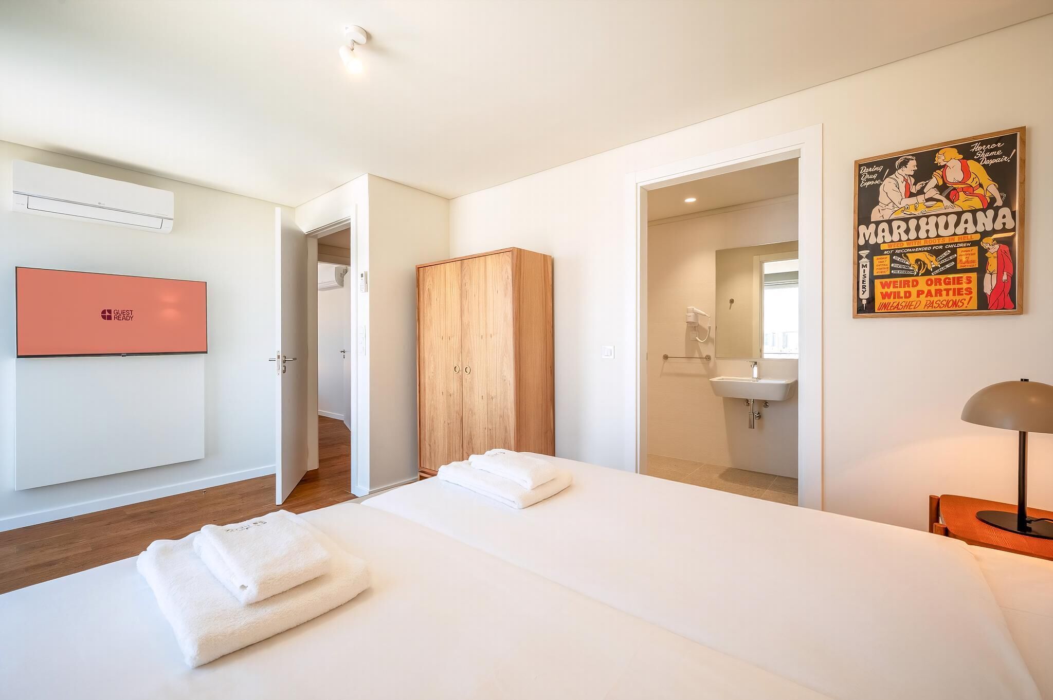 A hotel room with a bed, a wooden wardrobe, a bathroom with a sink, and a poster on the wall.