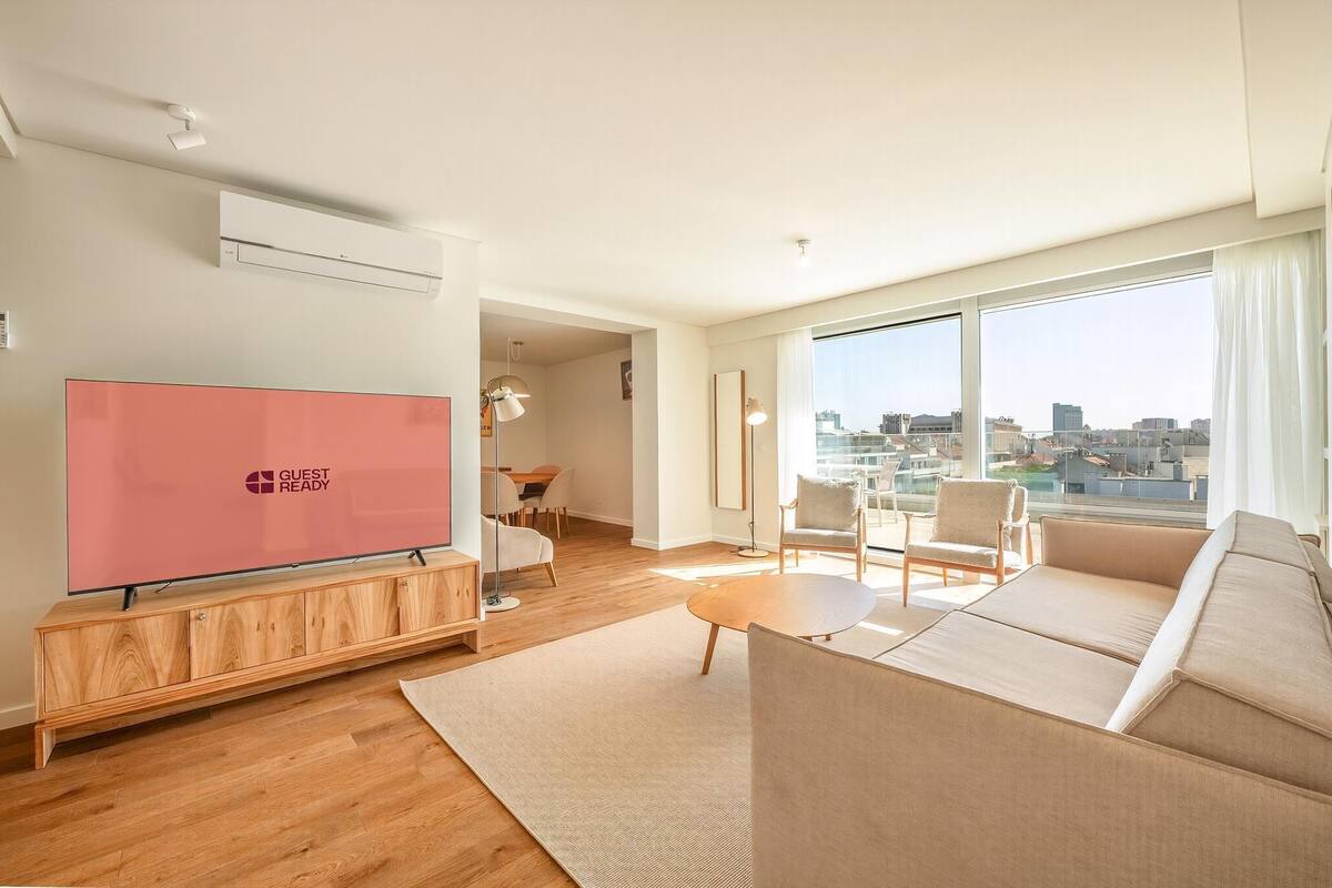 A modern living room with a flat-screen TV, a sofa, a coffee table, and a large window with a city view.