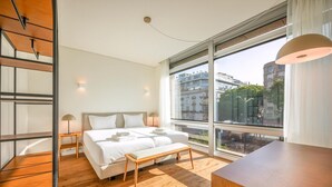 A modern hotel room with a large bed, a wooden bench, a desk, and a view of the city through large windows. - Guestready - Republic Apartments (Lisbon)
