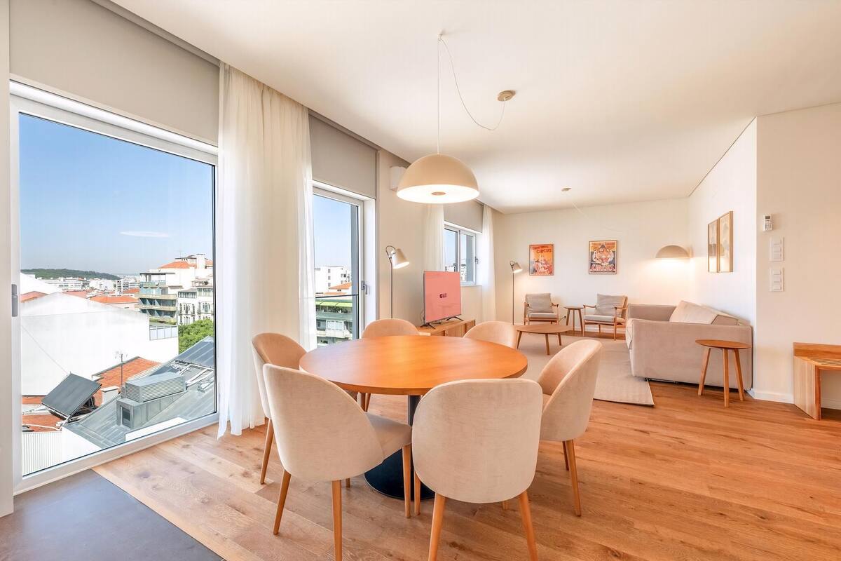 A modern living room with a round wooden table, beige chairs, a sofa, and a large window offering a city view.