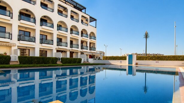 Een modern hotel met een zwembad, meerdere balkons en een heldere lucht.