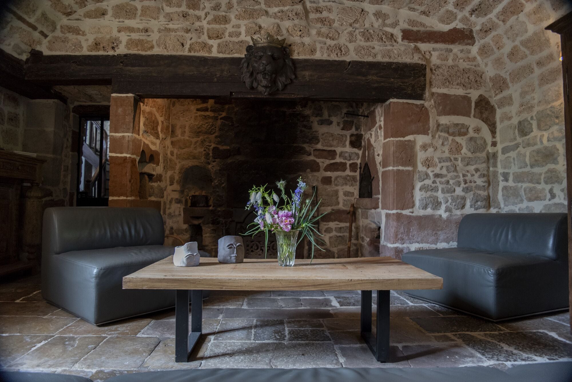 A stone-walled room with a fireplace, two leather sofas, a wooden table with a vase of flowers, and a crown above the fireplace.
