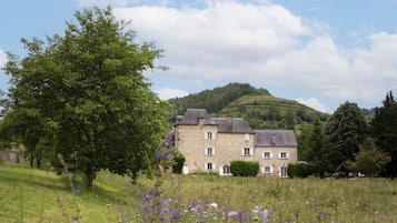 A stone building with a symmetrical facade, surrounded by a grassy field with purple flowers, under a partly cloudy sky.