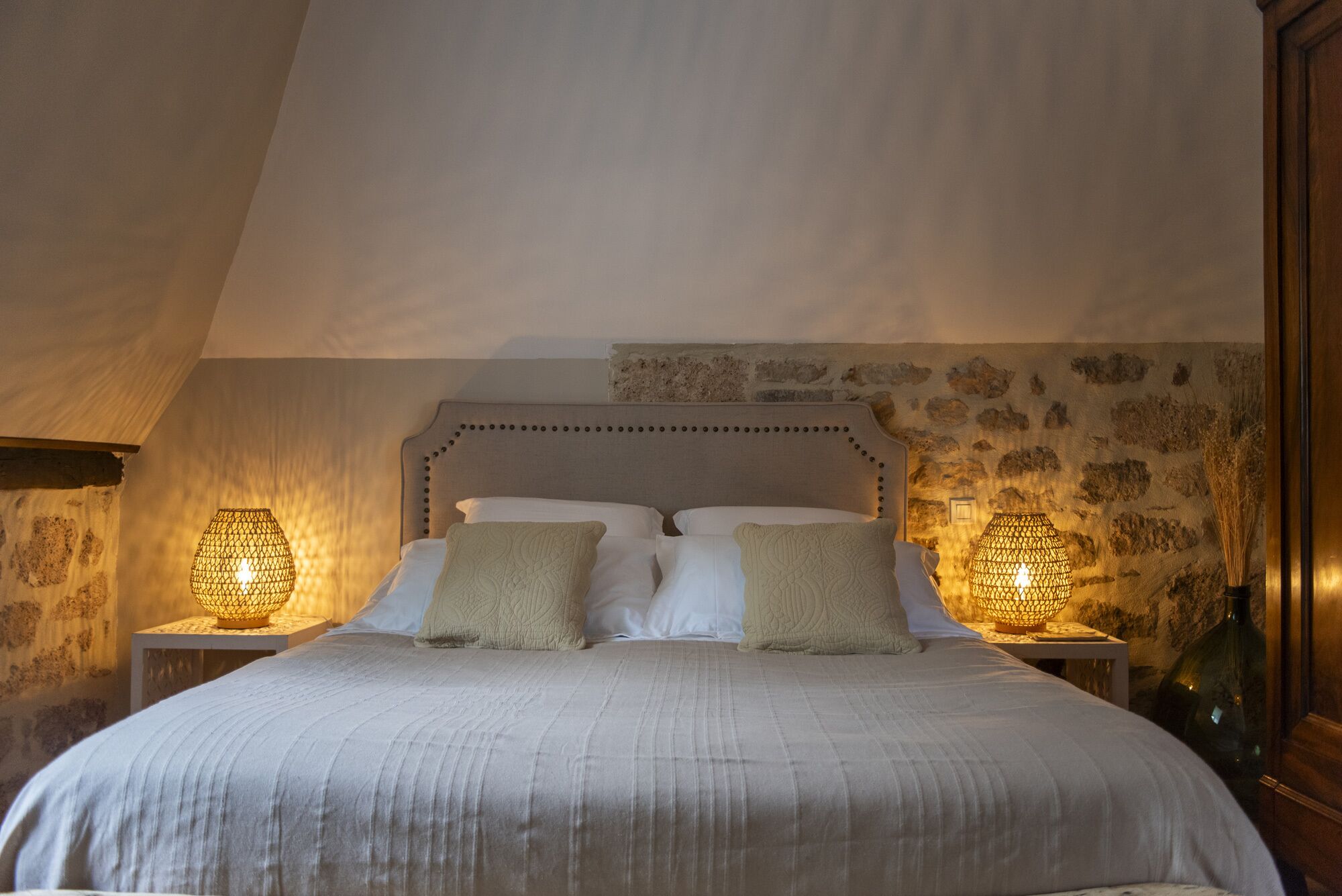 A bedroom with a large bed, two bedside lamps, and a stone wall.