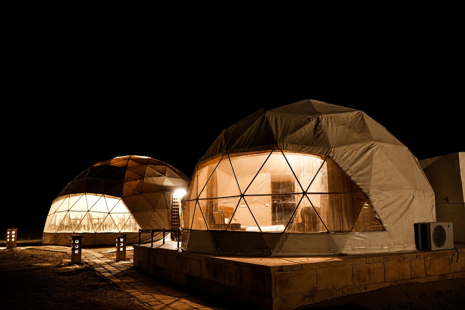 Two dome-shaped yurts with a warm interior light, a stone pathway, and a small outdoor area with a heater.