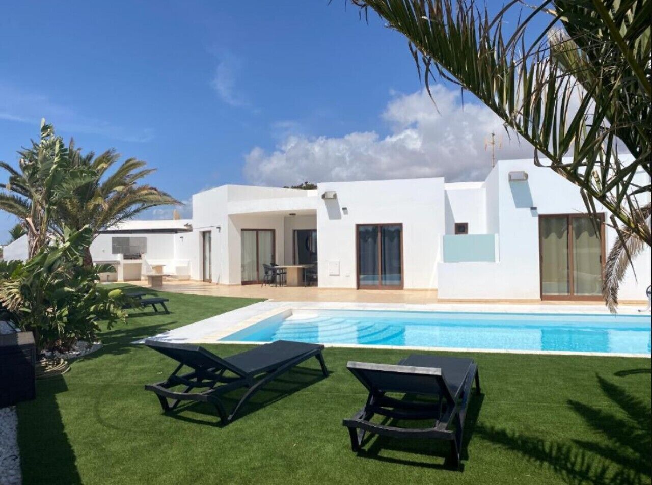 A modern house with a swimming pool, two lounge chairs, and a well-maintained lawn.