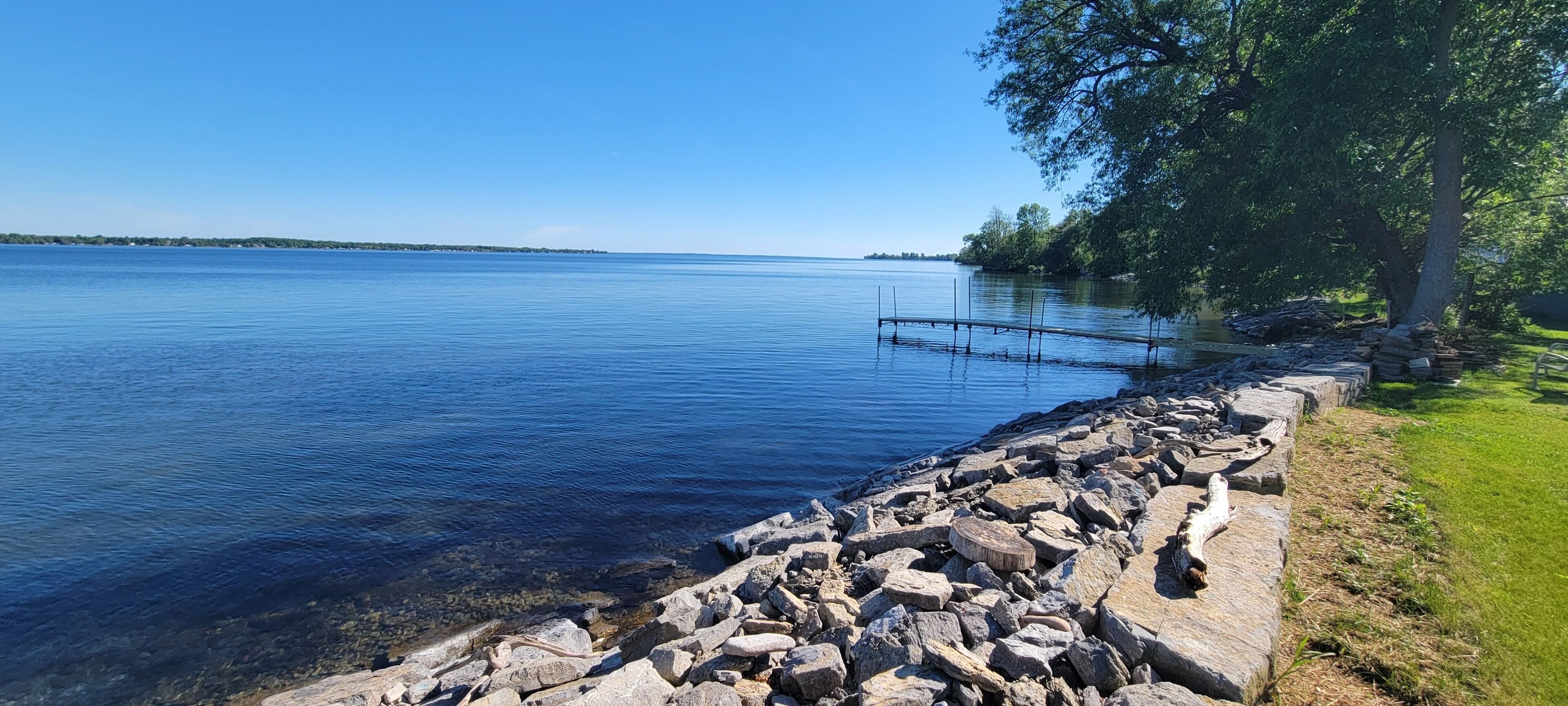 A calm lake with a rocky shoreline, a dock extending into the water, and a grassy area with trees.