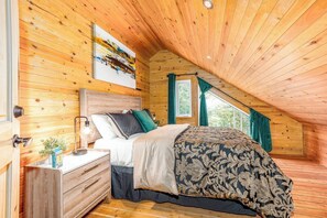 Um quarto aconchegante com teto inclinado, paredes de madeira, uma cama com colcha estampada, uma mesinha de cabeceira com abajur e uma janela com cortinas.