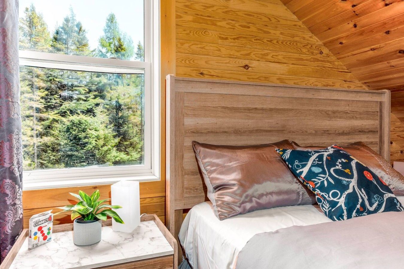 A wooden cabin room with a bed, a nightstand with a plant, and a window with a forest view.