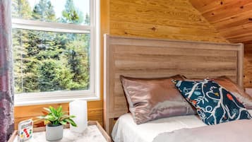 A wooden cabin room with a bed, a nightstand with a plant, and a window with a forest view.