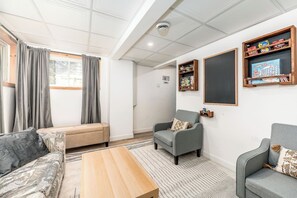 Quarto com sofá, poltrona e uma pequena mesa. Cortinas, estante e um quadro negro na parede.
