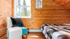 Um quarto de cabana aconchegante com paredes de madeira, janela com vista para árvores, cama com manta estampada, cadeira com travesseiro e quadro na parede.
