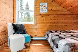 A cozy cabin room with wooden walls, a window with a view of trees, a bed with a patterned blanket, a chair with a pillow, and a framed picture on the wall.