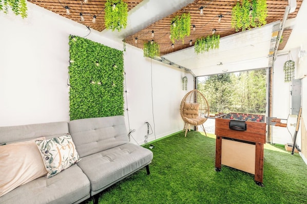 A room with a grey sofa, a foosball table, a hanging chair, and a green wall with plants.
