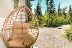 Une chaise en osier avec un coussin appuyé contre un mur, un chemin de gravier et une zone boisée avec de grands arbres.