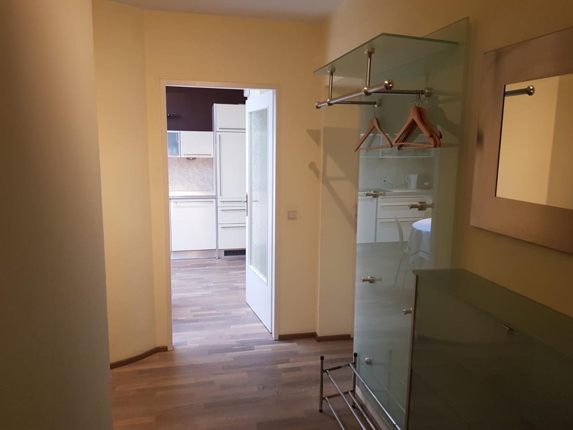 A hallway with a wooden floor leading to a kitchen area with white cabinets and a glass cabinet with a hanging rack.