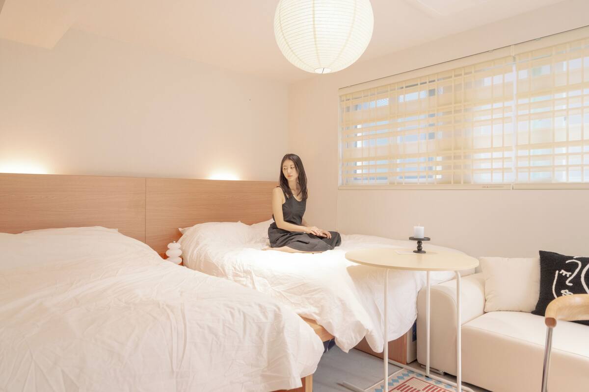 Habitación de hotel con dos camas, una mesita redonda pequeña, un sofá y un ventanal grande con persianas.