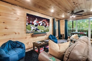 A wooden interior with a television displaying a football game, a sofa, a blue bean bag, and a table with magazines. - New! 3BR w/Hot Tub*Game Room*Fireplace*Privacy! (Pigeon Forge)