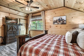 A wooden cabin interior with a bed, a dresser, a ceiling fan, and a window with a view of trees. - New! 3BR w/Hot Tub*Game Room*Fireplace*Privacy! (Pigeon Forge)