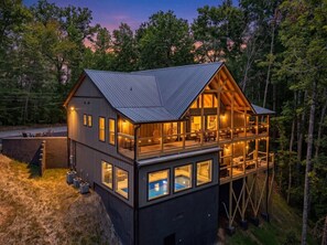 A two-story cabin with a large balcony, a pool, and a forested backdrop. - New! Loaded Luxe 4BR w/Pool*Hot Tub*Sauna*Views! (Sevierville)