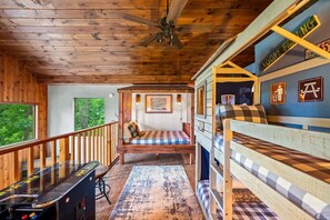 Een blokhutinterieur met houten plafond, een stapelbed, een bureau met computer en uitzicht op de natuur.