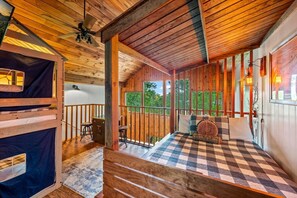 A wooden cabin interior with a checkered floor, a bed with a wooden headboard, a desk, and a balcony with a view of greenery. - 2BR w/Treehouse! Hot Tub*Arcade*Fireplace*Privacy! (Sevierville)