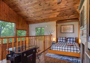 A cabin with a wooden interior, a bed with checkered bedding, a desk with a computer, and a window overlooking trees. - 2BR w/Treehouse! Hot Tub*Arcade*Fireplace*Privacy! (Sevierville)