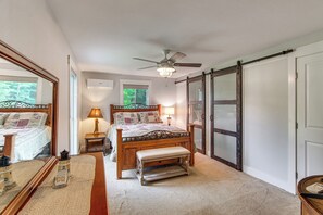 Quarto com cama, banqueta, mesa de cabeceira, abajur, ventilador de teto e um espelho grande.