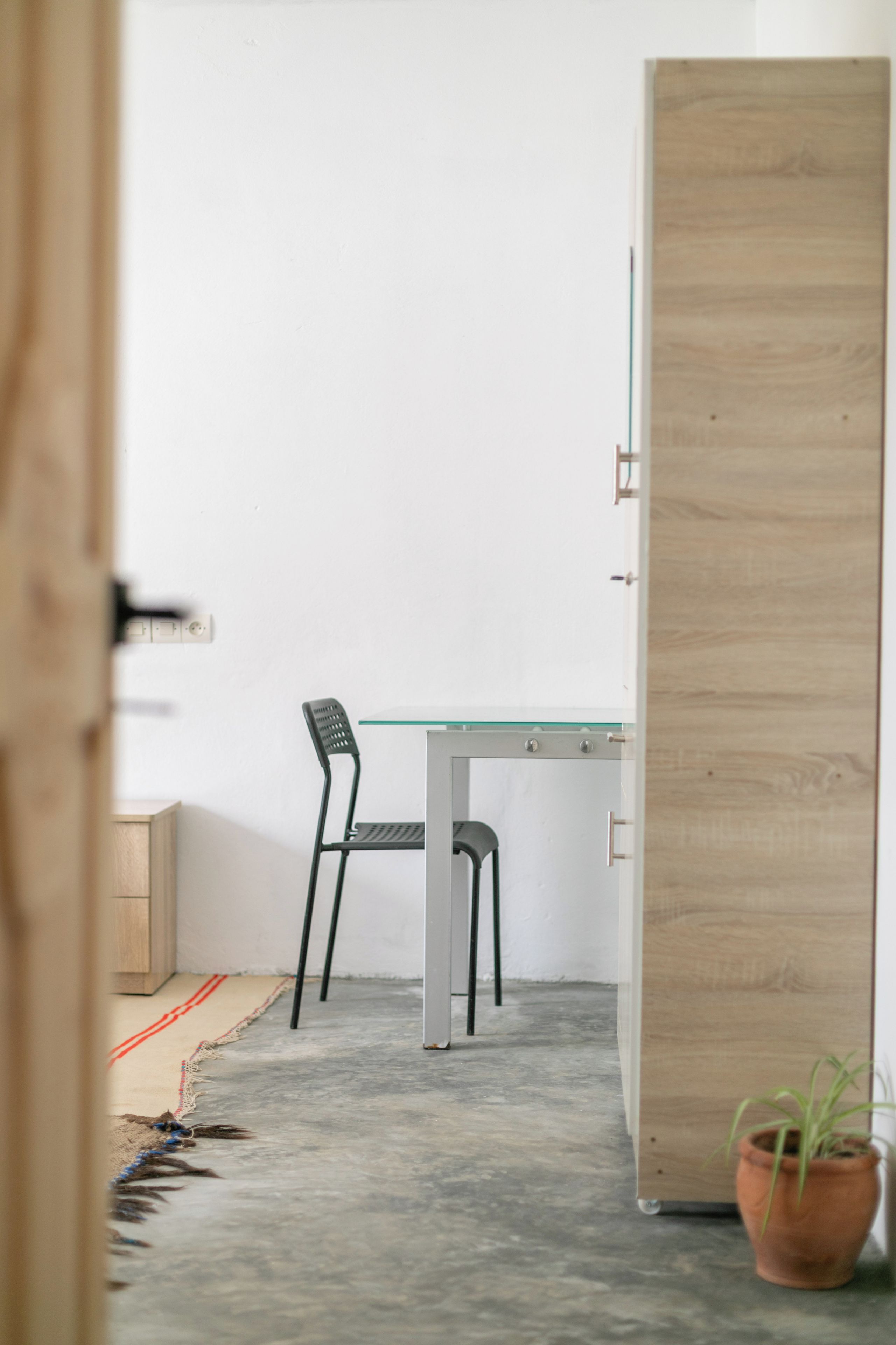 Quarto com escrivaninha de tampo de vidro, duas cadeiras, planta em vaso e porta de madeira.