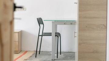Quarto com escrivaninha de tampo de vidro, duas cadeiras, planta em vaso e porta de madeira.